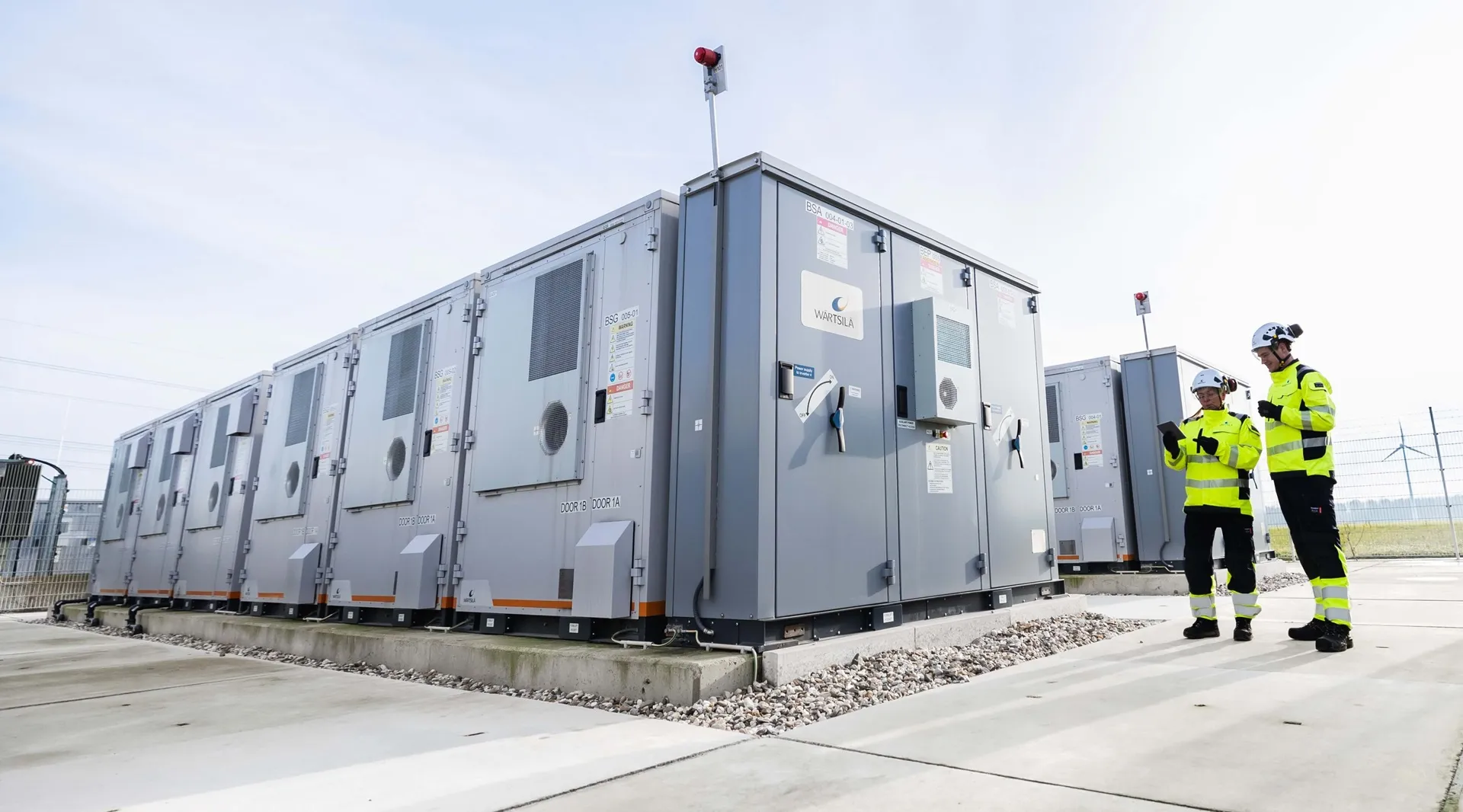 A row of Wärtsilä battery energy storage enclosures on a concrete pad, two engineers checking a tablet alongside them