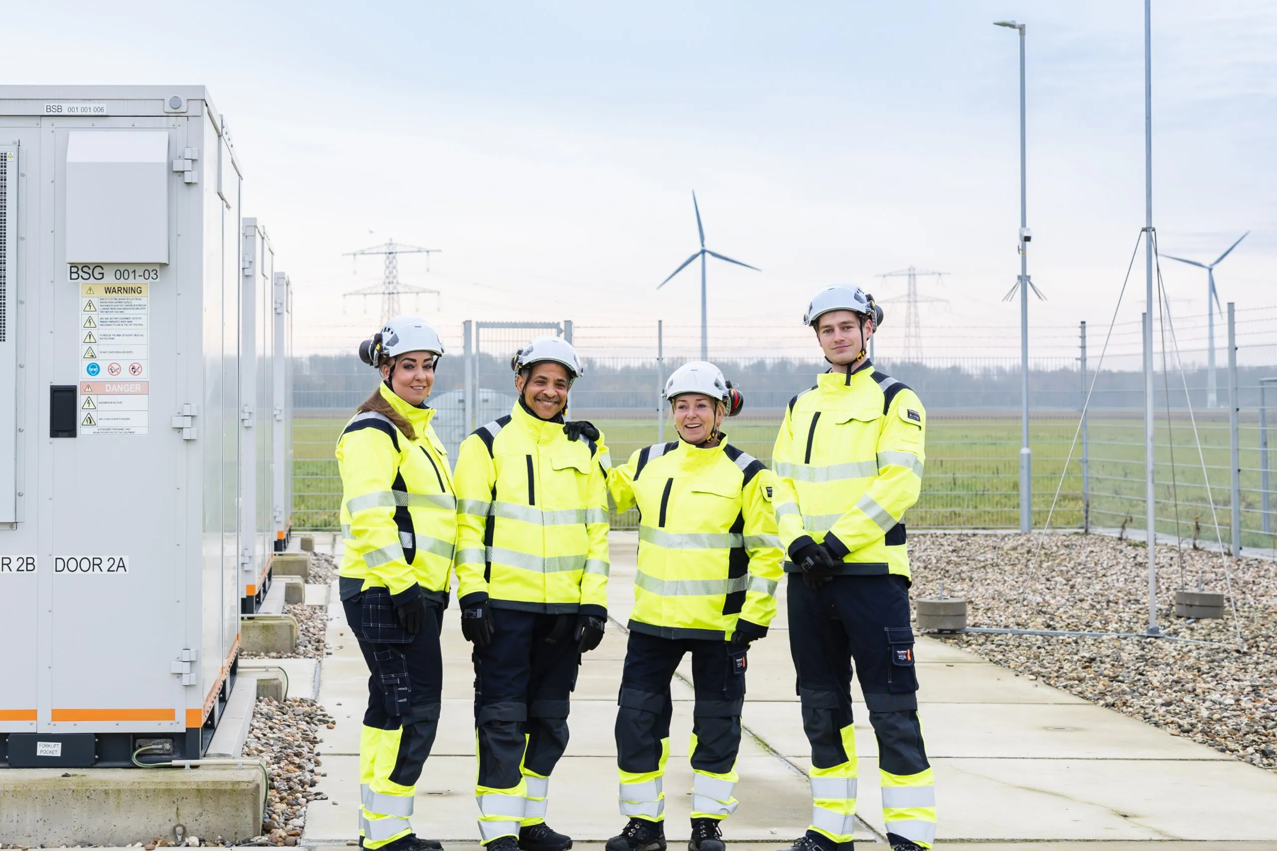 Group of technicians in hard helmet and high visibility gear with wind mills in background.