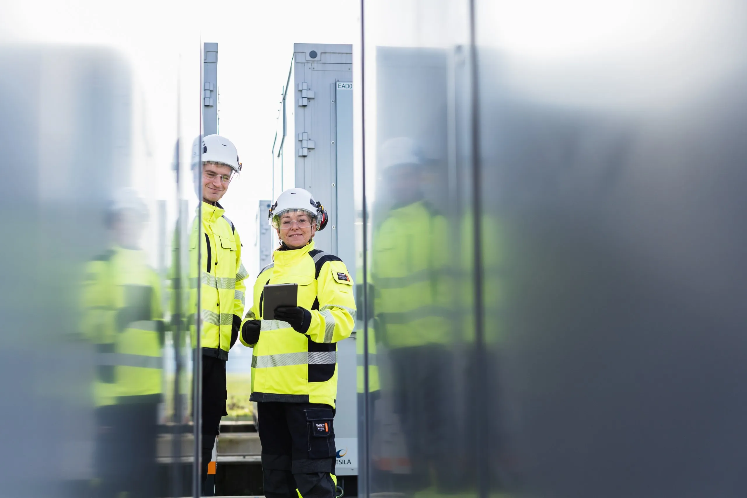 Two technicians in high-visibility gear and hard hats, one holding a tablet, standing between battery storage units.