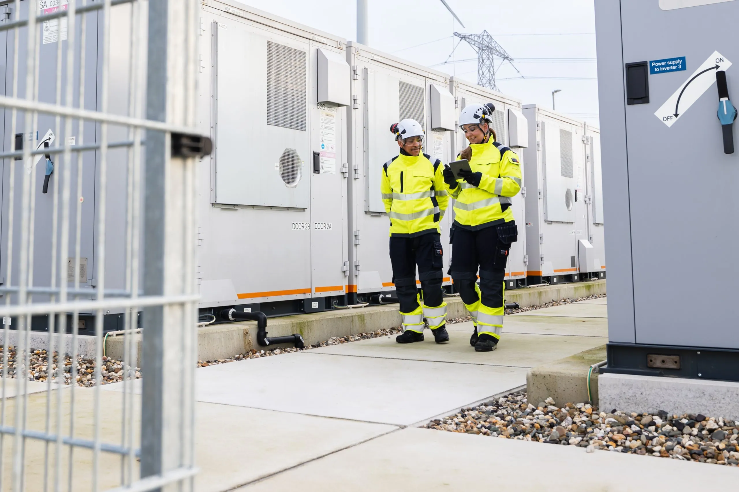 Two technicians in high-visibility gear and hard hats walking between storage units.