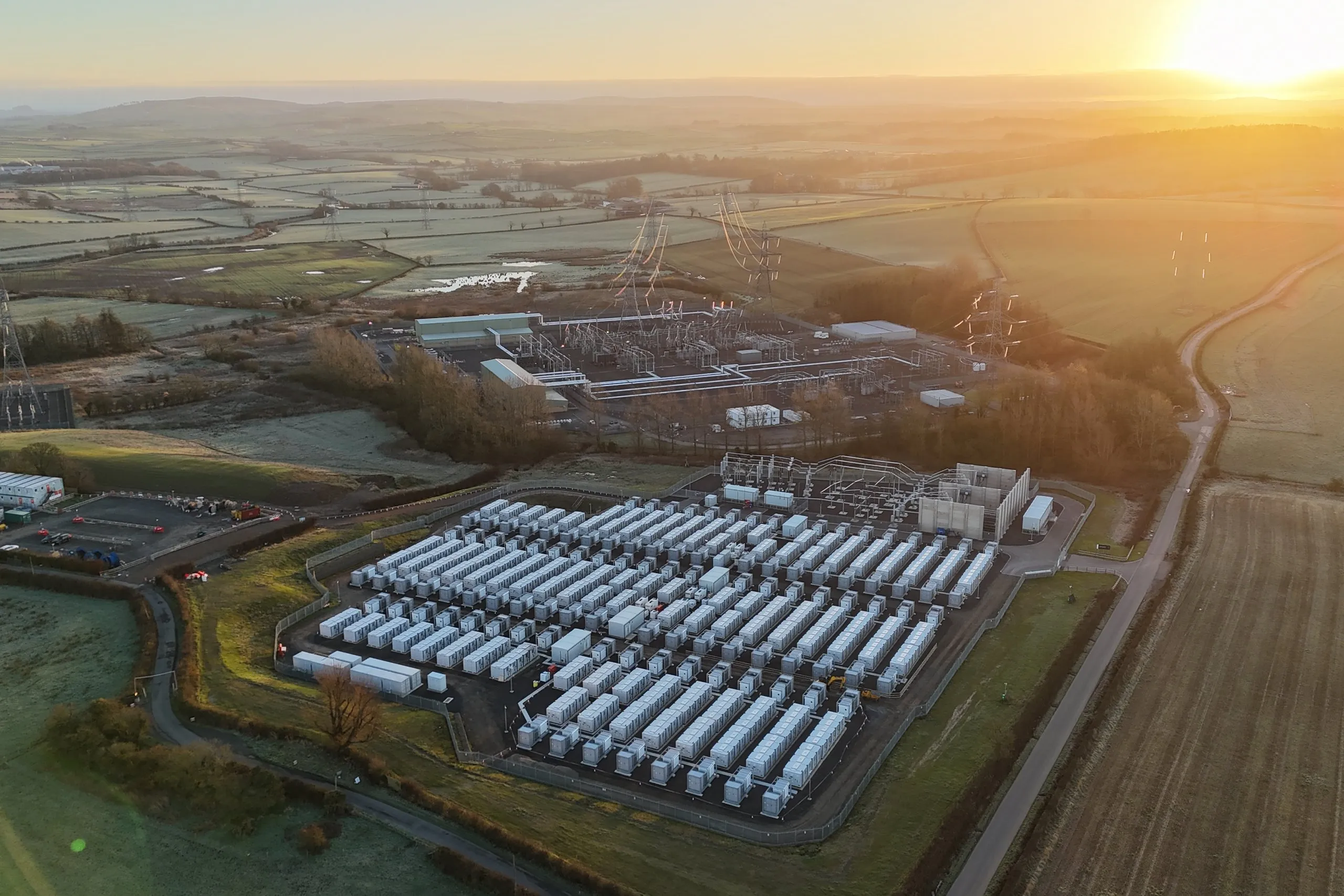 Aerial view of a large battery energy storage facility surrounded by green fields at sunset.