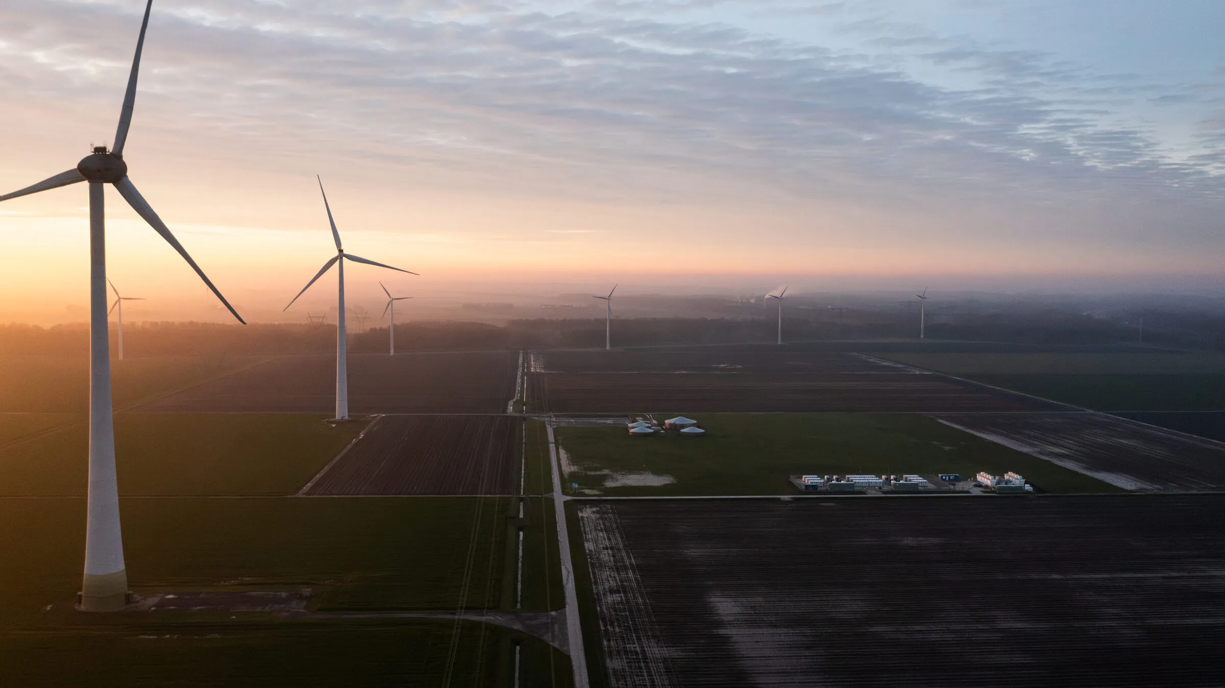 Wind farm at sunrise seen from the air, turbines over ploughed fields with a battery storage compound in the middle distance
